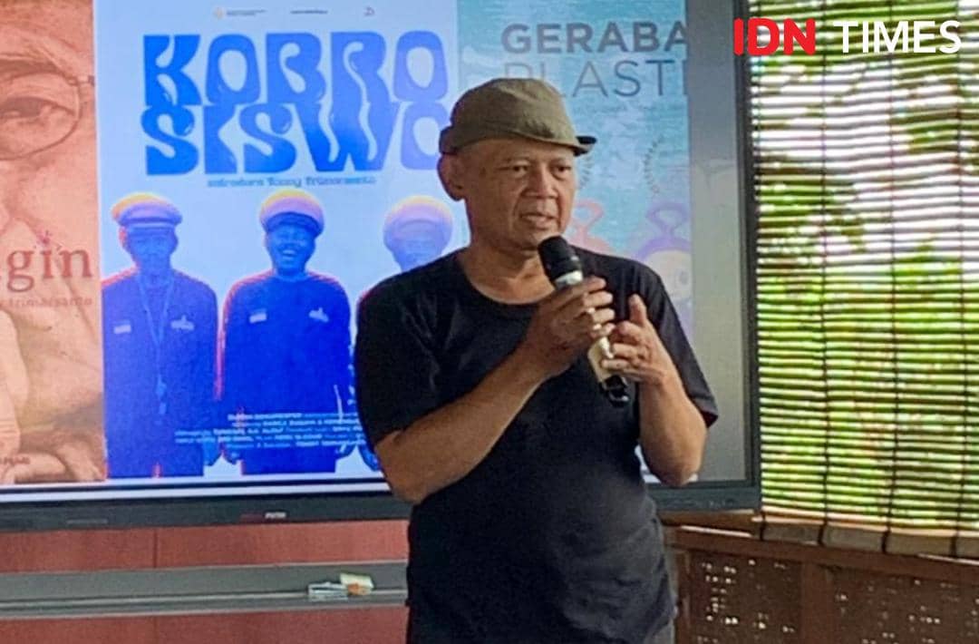 Seorang pria mengenakan kaus hitam dan topi abu-abu memegang mikrofon sambil berbicara di depan layar dengan poster film di latar belakang.