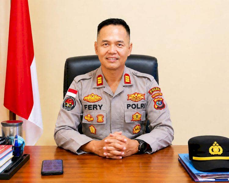 Seorang perwira polisi berpakaian dinas duduk di meja kerjanya dengan tangan terlipat, di samping topi dinas dan bendera Indonesia.