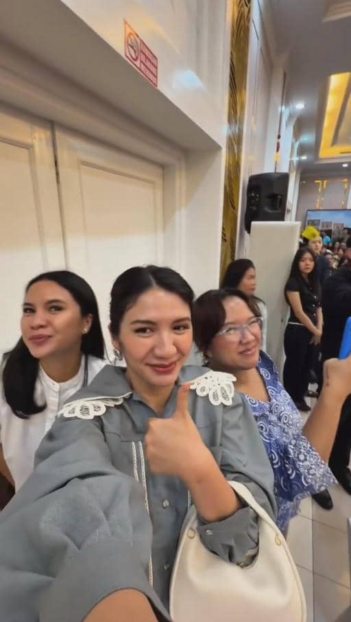 Seorang wanita tersenyum sambil mengacungkan jempol bersama dua wanita lain di dalam ruangan acara wisuda yang ramai.