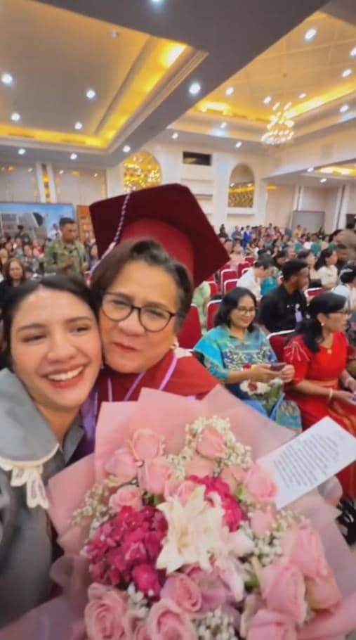 Dua perempuan tersenyum bahagia saat wisuda, salah satunya mengenakan toga merah dan memegang buket bunga berwarna pink di aula penuh tamu.