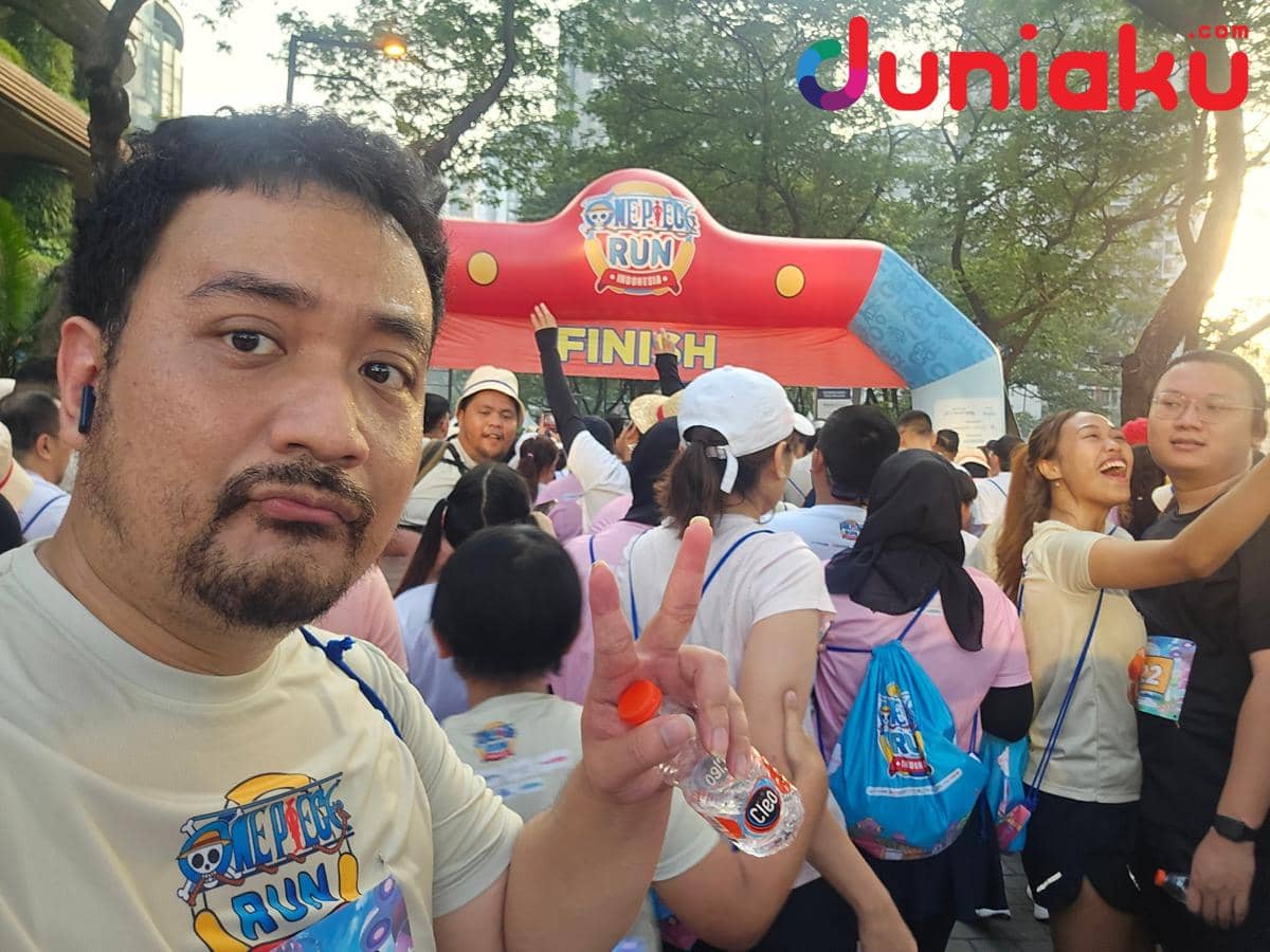 Seorang peserta berfoto selfie sambil menunjukkan tanda damai di depan gerbang finish acara lari ONE PIECE Run 2026 yang ramai oleh pelari lain.