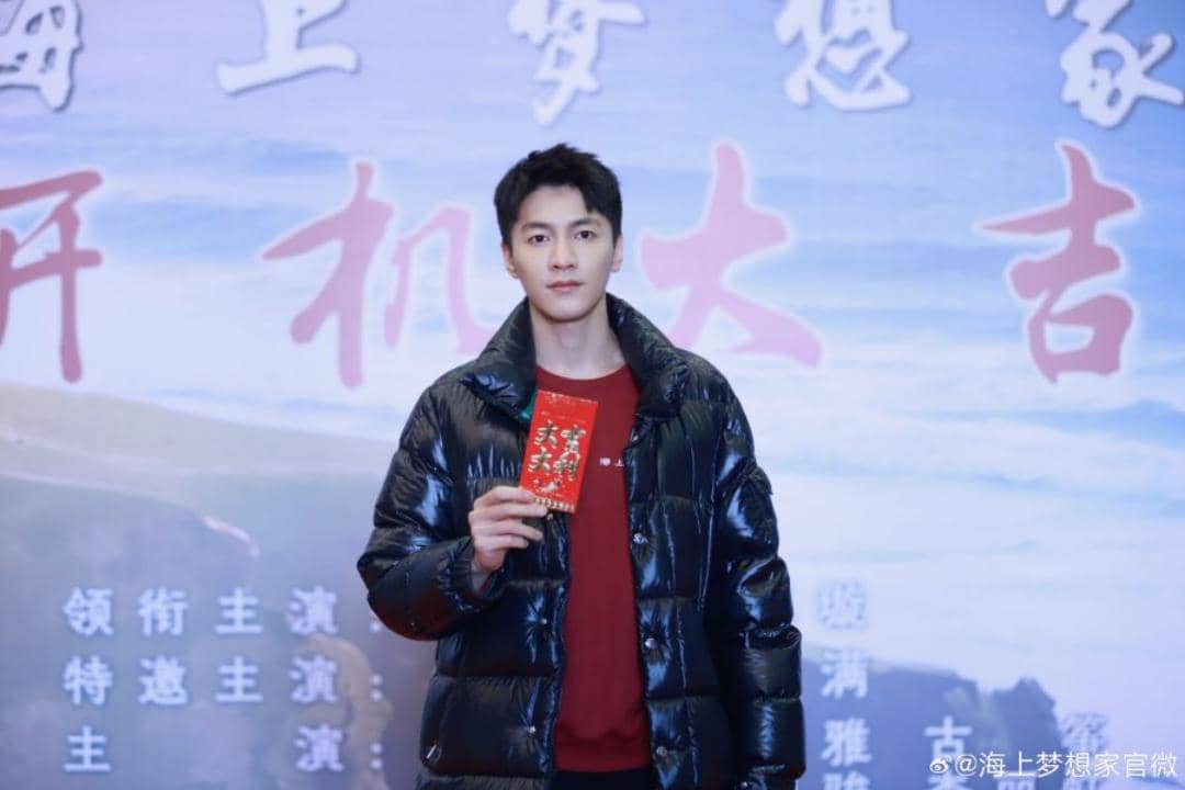 Seorang pria mengenakan jaket hitam memegang amplop merah di depan latar bertulisan Mandarin pada acara terkait Begin Again.