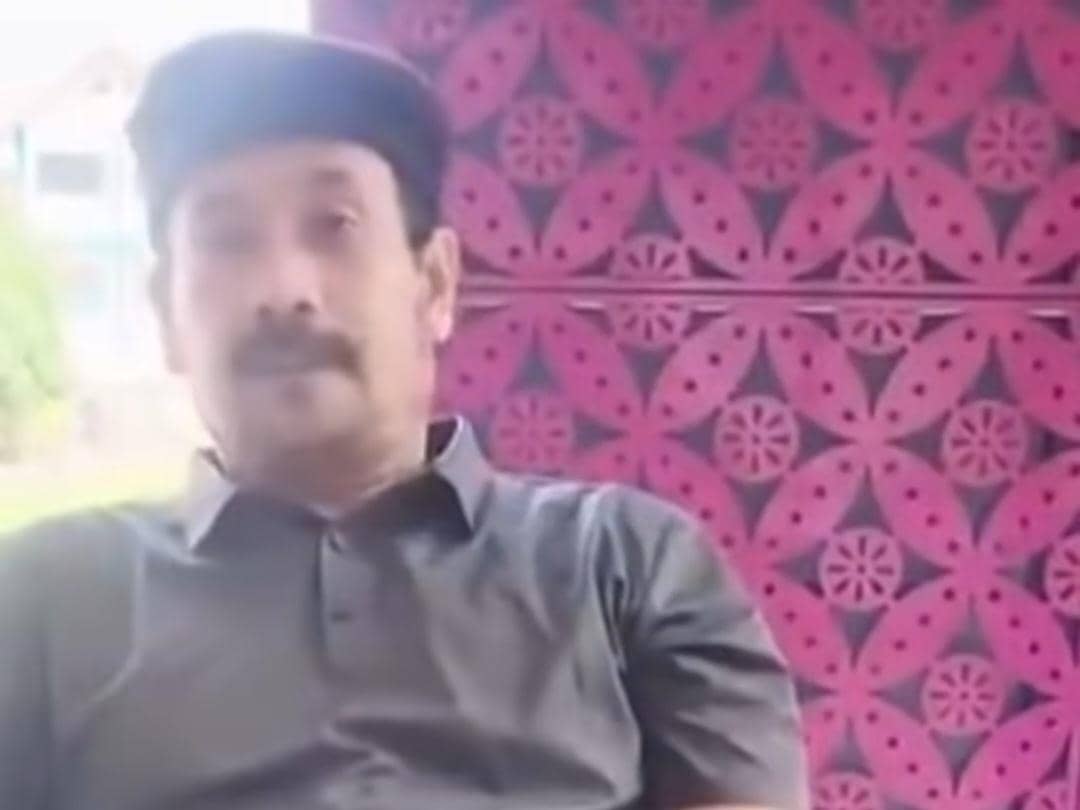 Seorang pria mengenakan kemeja abu-abu dan topi hitam duduk di depan dinding bermotif ungu dan merah muda dengan ekspresi tenang.