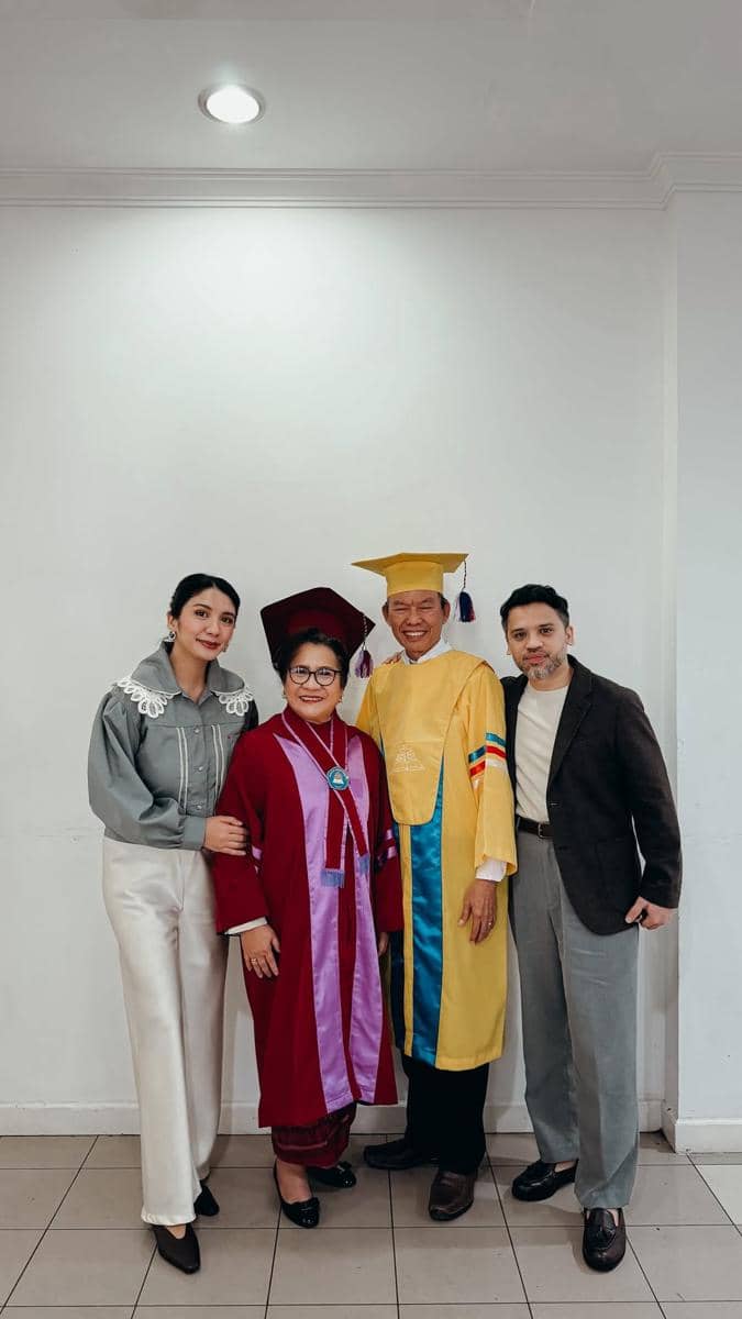 Empat orang berpose bersama di dalam ruangan, dua di antaranya mengenakan toga wisuda berwarna merah dan kuning dengan senyum bahagia.
