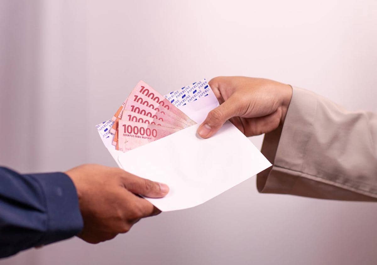Dua tangan sedang menyerahkan amplop putih berisi uang kertas seratus ribu rupiah di latar belakang polos berwarna terang.