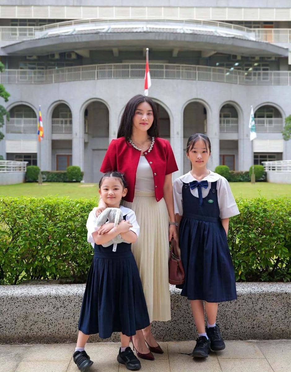 Seorang wanita mengenakan cardigan merah dan rok putih berdiri bersama dua anak perempuan berseragam sekolah biru tua di depan gedung bertingkat.