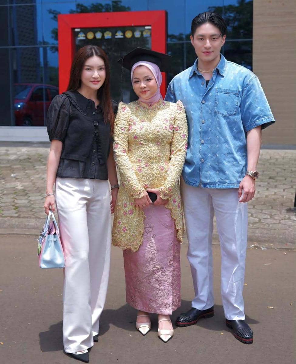 Tiga orang berpose bersama di luar ruangan, dua mengenakan celana putih dan satu memakai kebaya kuning dengan hijab serta toga wisuda.