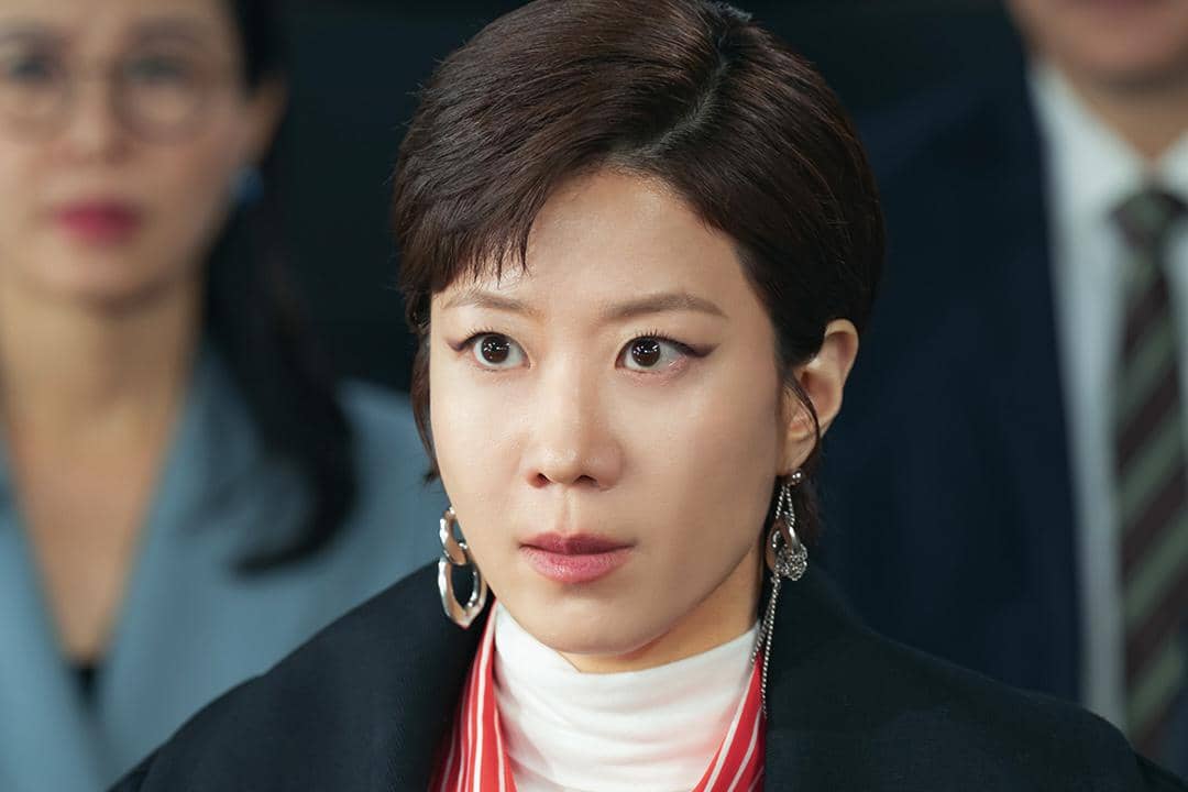 Seorang wanita berambut pendek mengenakan anting besar dan pakaian formal dengan ekspresi serius dalam adegan drama Korea Reborn Rookie.