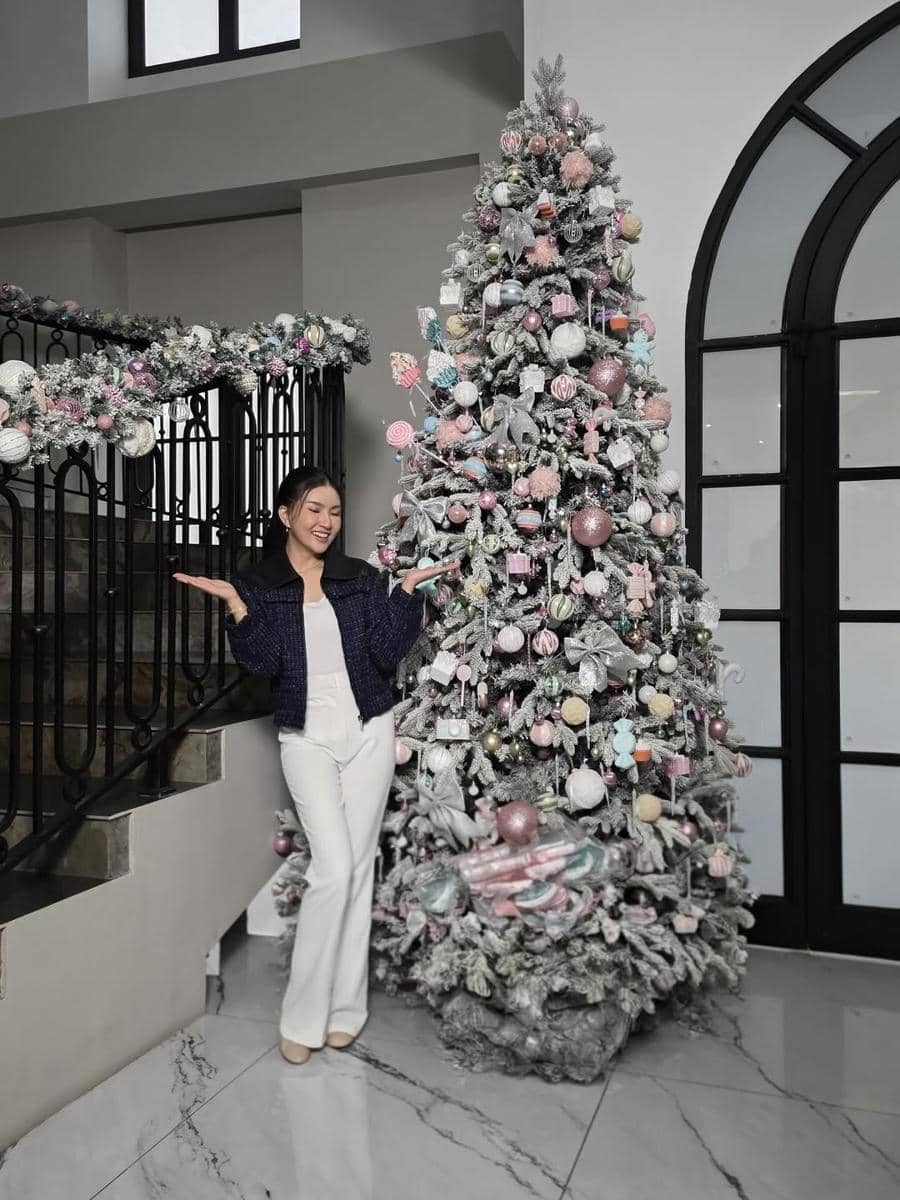 Seorang wanita berpose di samping pohon Natal besar yang dihiasi ornamen berwarna pastel di dalam ruangan bergaya modern.