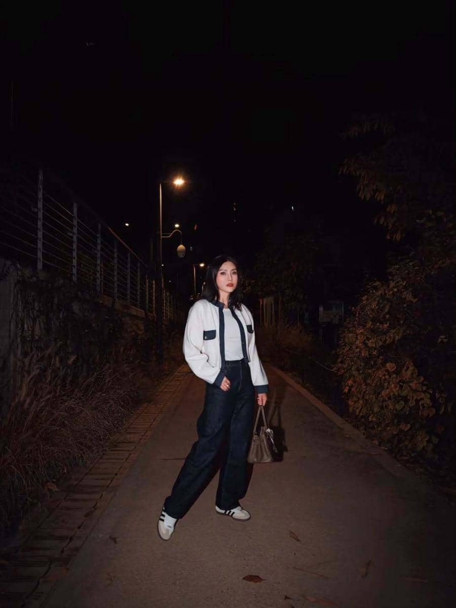Seorang wanita bergaya kasual mengenakan jaket putih dan celana panjang gelap sambil membawa tas tangan di jalan malam yang diterangi lampu jalan.