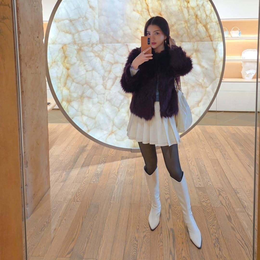 Seorang wanita berpose di depan cermin mengenakan jaket bulu ungu tua, rok putih, dan sepatu bot putih tinggi di ruangan modern.