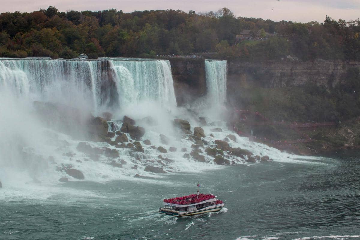 Perahu wisata penuh penumpang mendekati air terjun besar Niagara Falls dengan semburan air dan tebing hijau di latar belakang.