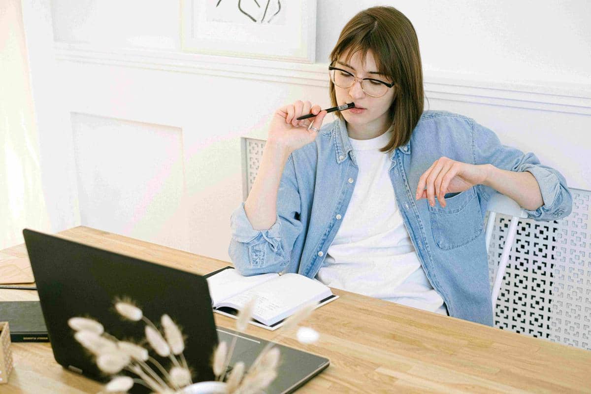 Seorang perempuan duduk di meja kerja dengan laptop terbuka dan buku catatan, tampak berpikir sambil memegang pena di bibirnya.