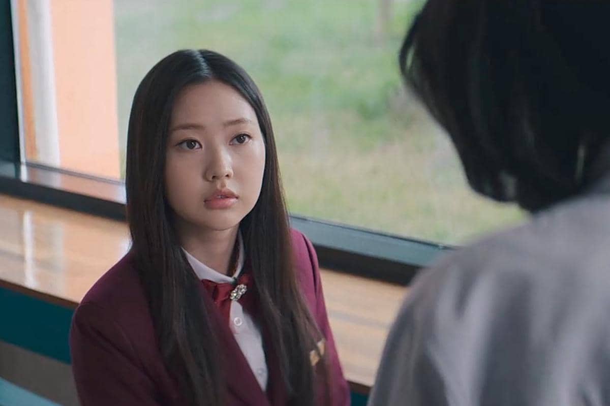 Seorang siswi berseragam merah duduk di dekat jendela sambil menatap seseorang dalam cuplikan drama Teach You a Lesson.