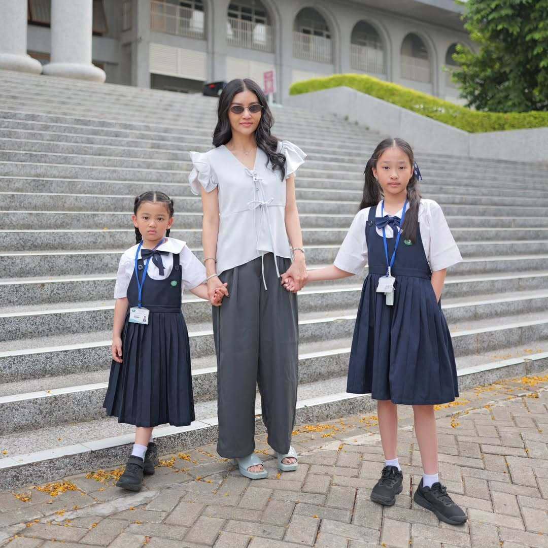 Seorang wanita mengenakan blus putih dan celana abu-abu berdiri di tangga sambil memegang tangan dua anak berseragam sekolah.