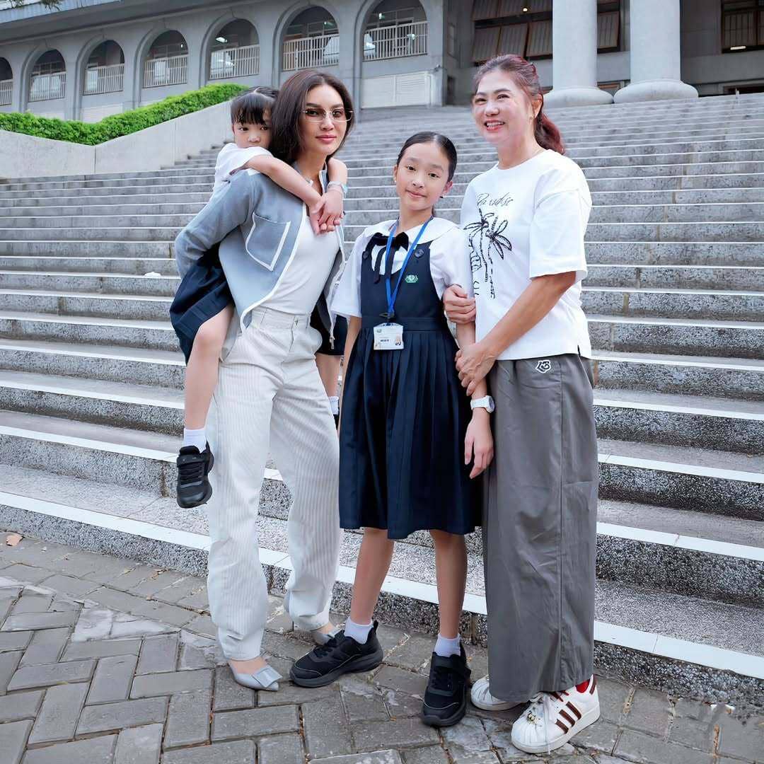 Seorang wanita mengenakan outfit putih bergaya kasual berpose bersama dua anak dan seorang wanita lain di depan tangga besar luar ruangan.