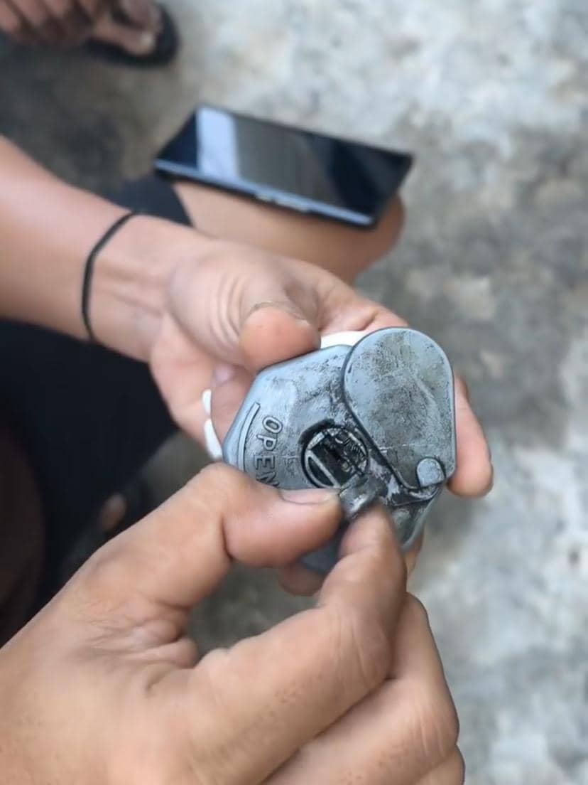 Seseorang memegang gembok logam berwarna abu-abu yang tampak rusak sambil mencoba membuka bagian kuncinya di area luar ruangan.