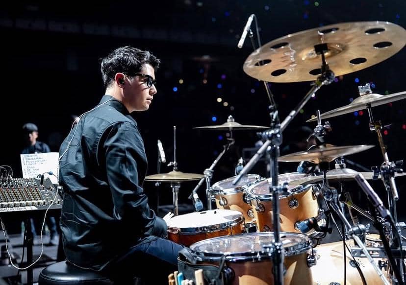 Seorang pria mengenakan pakaian hitam sedang memainkan drum di atas panggung konser dengan pencahayaan biru dan penonton di latar belakang.