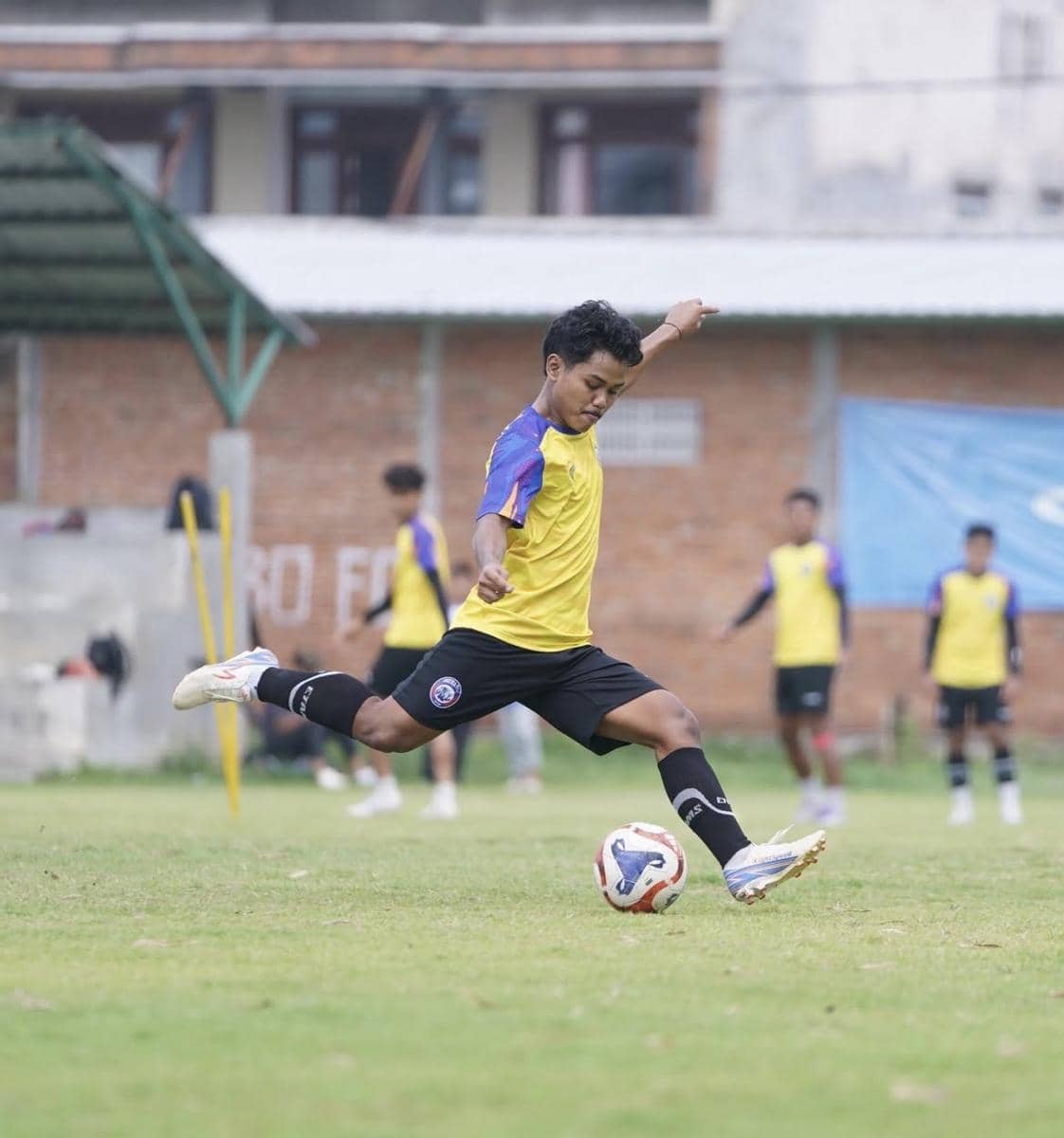 Seorang pemain Arema FC mengenakan jersey latihan kuning dan menendang bola di lapangan dengan beberapa rekan setim di latar belakang.