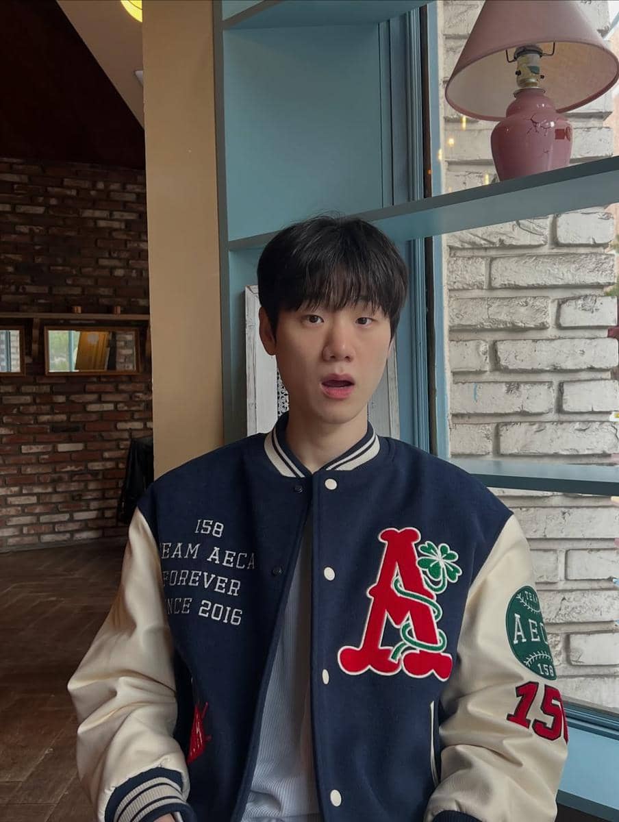 Seorang pria berjaket varsity biru krem duduk di dekat jendela dengan ekspresi terkejut di dalam ruangan bergaya kafe.