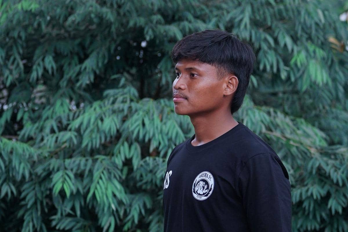 Seorang pemain Arema FC mengenakan kaus hitam dengan logo klub di dada, berdiri di depan latar pepohonan hijau.