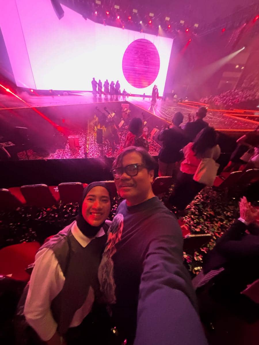 Dua orang berfoto selfie di antara penonton konser dengan panggung besar bercahaya merah muda dan pertunjukan sedang berlangsung di depan mereka.