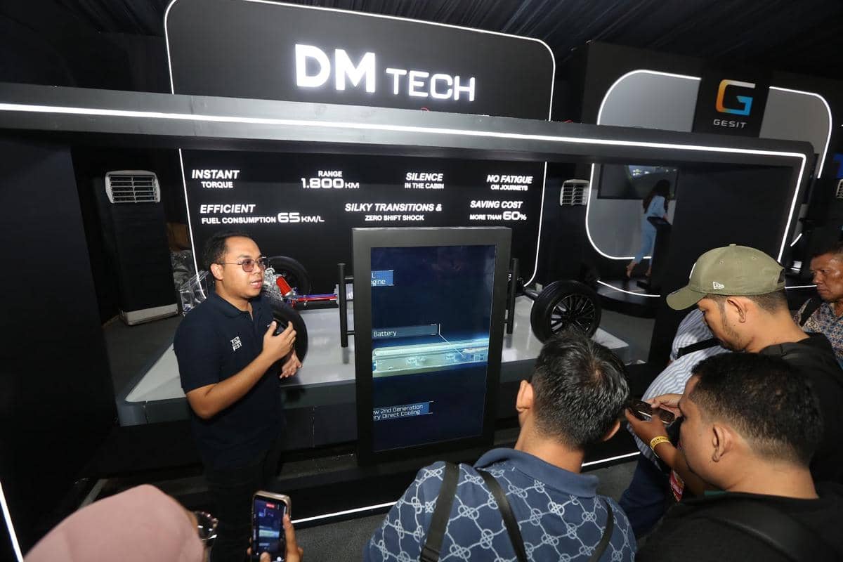 Seorang presenter menjelaskan teknologi Dual Mode BYD di depan layar interaktif kepada beberapa pengunjung di sebuah pameran otomotif.