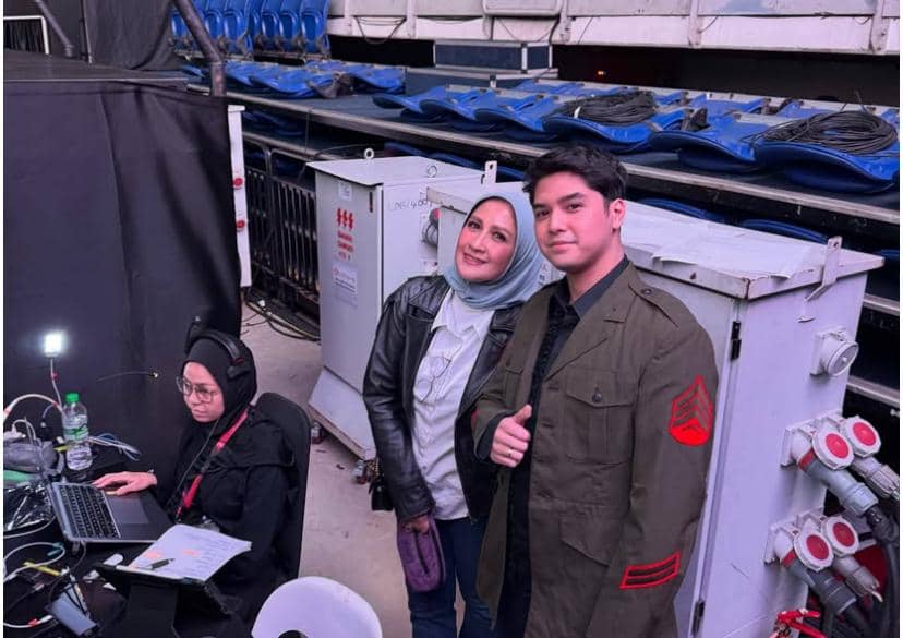 Seorang pria berjaket hijau berpose bersama seorang wanita berhijab di area belakang panggung konser dengan peralatan teknis di sekitarnya.