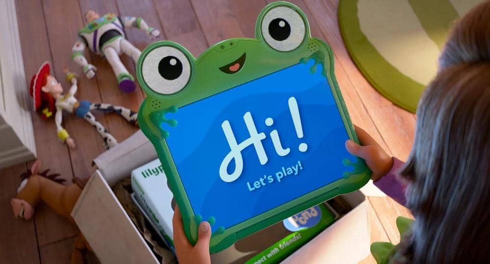 Seorang anak memegang tablet berbentuk katak dengan layar bertuliskan 'Hi! Let's play!' di dekat mainan karakter Toy Story di lantai kayu.