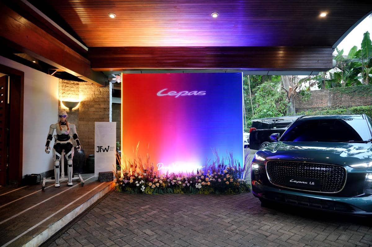 Mobil Lepas L8 dipajang di area acara JFW 2027 dengan latar belakang papan berwarna gradasi dan robot AiMOGA di sisi kiri.