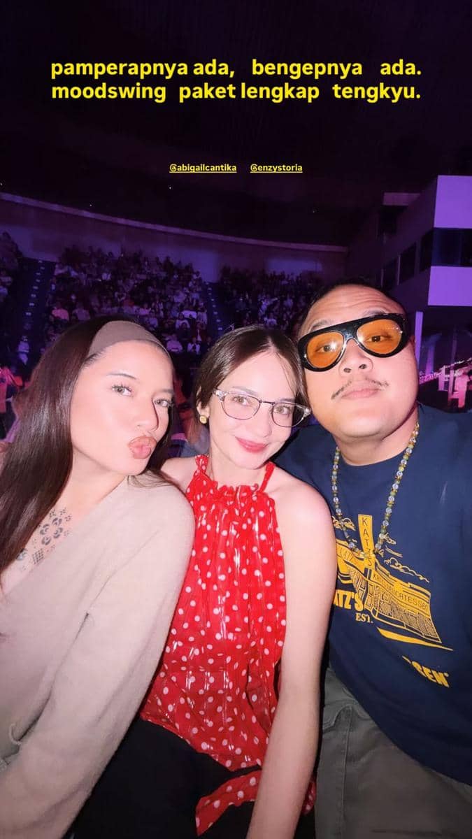 Tiga orang berpose bersama di dalam arena konser dengan pencahayaan ungu, tampak penonton memenuhi tribun di belakang mereka.
