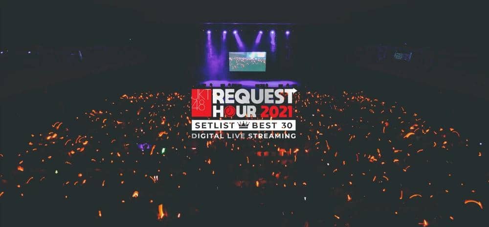 Panggung konser JKT48 Request Hour 2021 dengan pencahayaan ungu dan penonton memegang lightstick oranye di area gelap.