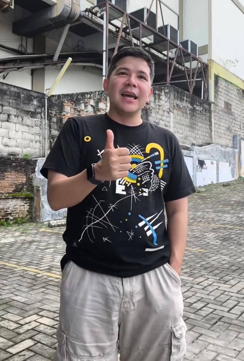 Seorang pria mengenakan kaus hitam bergambar warna-warni dan celana pendek krem berdiri di area luar dengan latar dinding bata dan bangunan industri.