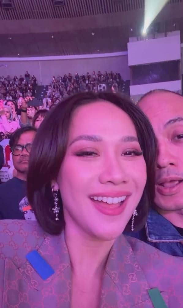 Seorang wanita tersenyum lebar saat menonton konser di arena besar yang dipenuhi penonton dengan pencahayaan ungu dan merah muda.