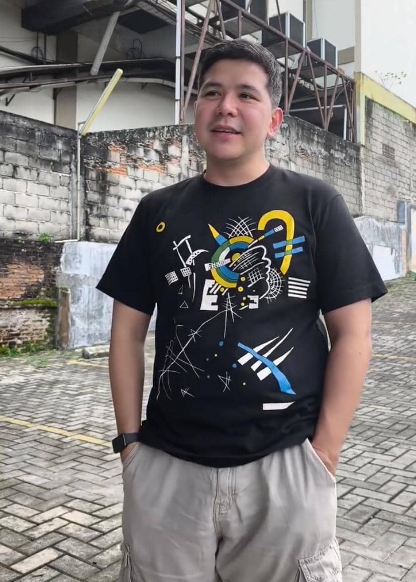 Seorang pria mengenakan kaus hitam bergambar abstrak warna-warni dan celana pendek krem berdiri di area luar dengan dinding bata di belakangnya.