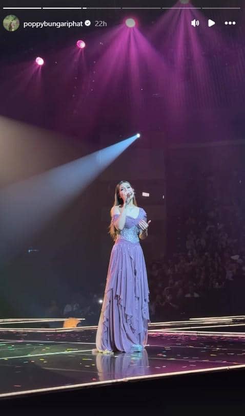 Seorang penyanyi mengenakan gaun ungu tampil di atas panggung konser dengan pencahayaan sorot berwarna ungu dan penonton di latar belakang.