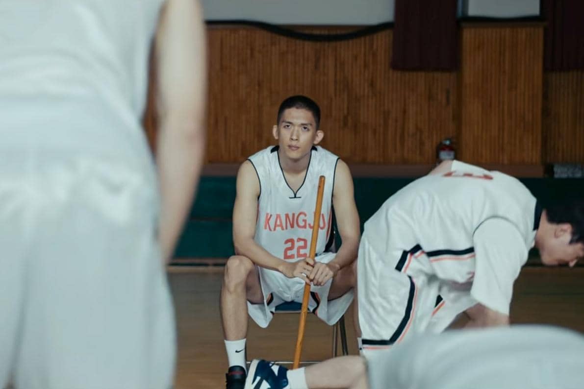 Pemain drama Korea Teach You a Lesson mengenakan seragam basket bertuliskan Kangjo duduk di kursi di dalam gedung olahraga.