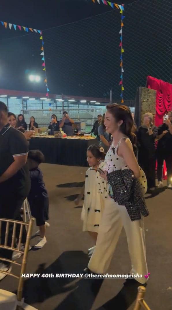 Seorang wanita tersenyum berjalan bersama anak kecil di acara ulang tahun ke-40 yang berlangsung di area luar ruangan pada malam hari.