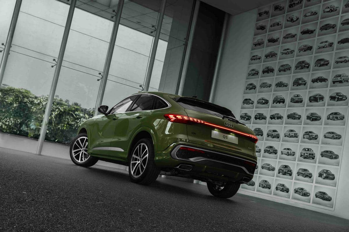 Audi Q5 Sportback berwarna hijau tampak dari belakang di dalam ruangan dengan dinding kaca dan deretan foto mobil di latar belakang.