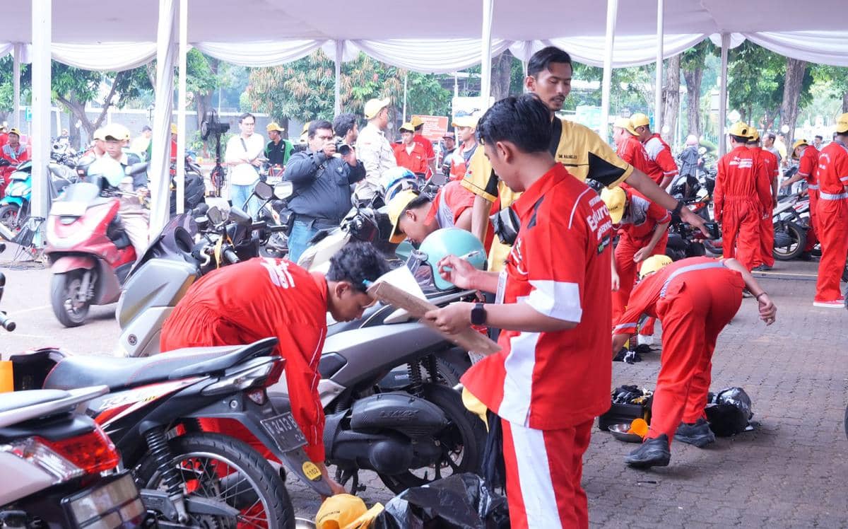 Siswa SMK mengenakan seragam merah sedang mengganti oli motor di area terbuka dengan pengawasan instruktur.