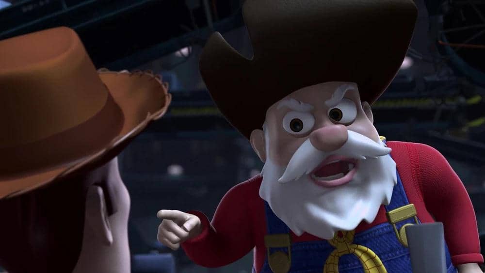 Karakter mainan koboi tua berjanggut putih mengenakan topi cokelat dan pakaian merah sedang berbicara dengan karakter lain di Toy Story 2.