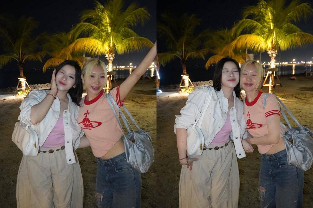 Dua perempuan berpose ceria di pantai pada malam hari dengan latar pohon kelapa dan lampu kota di kejauhan.
