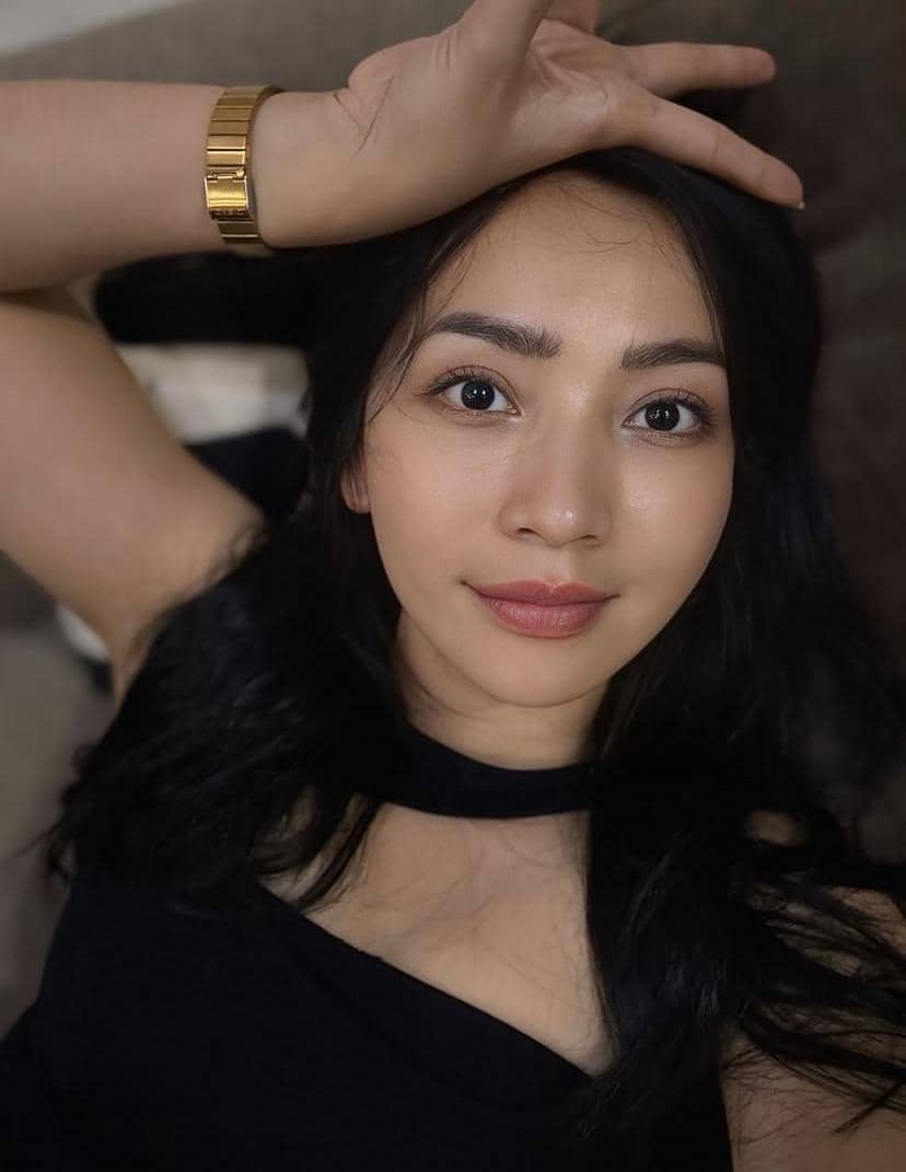 Seorang perempuan berambut hitam panjang mengenakan pakaian hitam berpose santai sambil mengangkat tangan di atas kepala.