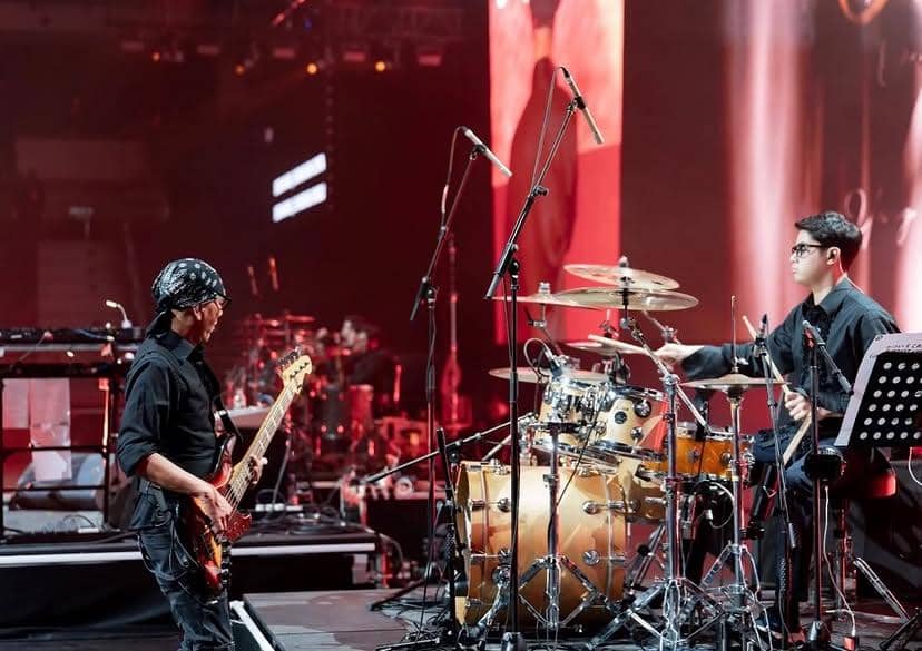 Seorang drummer muda tampil di atas panggung bersama pemain bass saat konser Dewa 19 di Kuala Lumpur dengan pencahayaan merah.