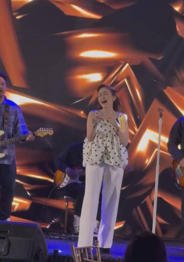 Seorang penyanyi wanita bernyanyi di atas panggung bersama band, dengan latar belakang lampu panggung berwarna oranye keemasan.