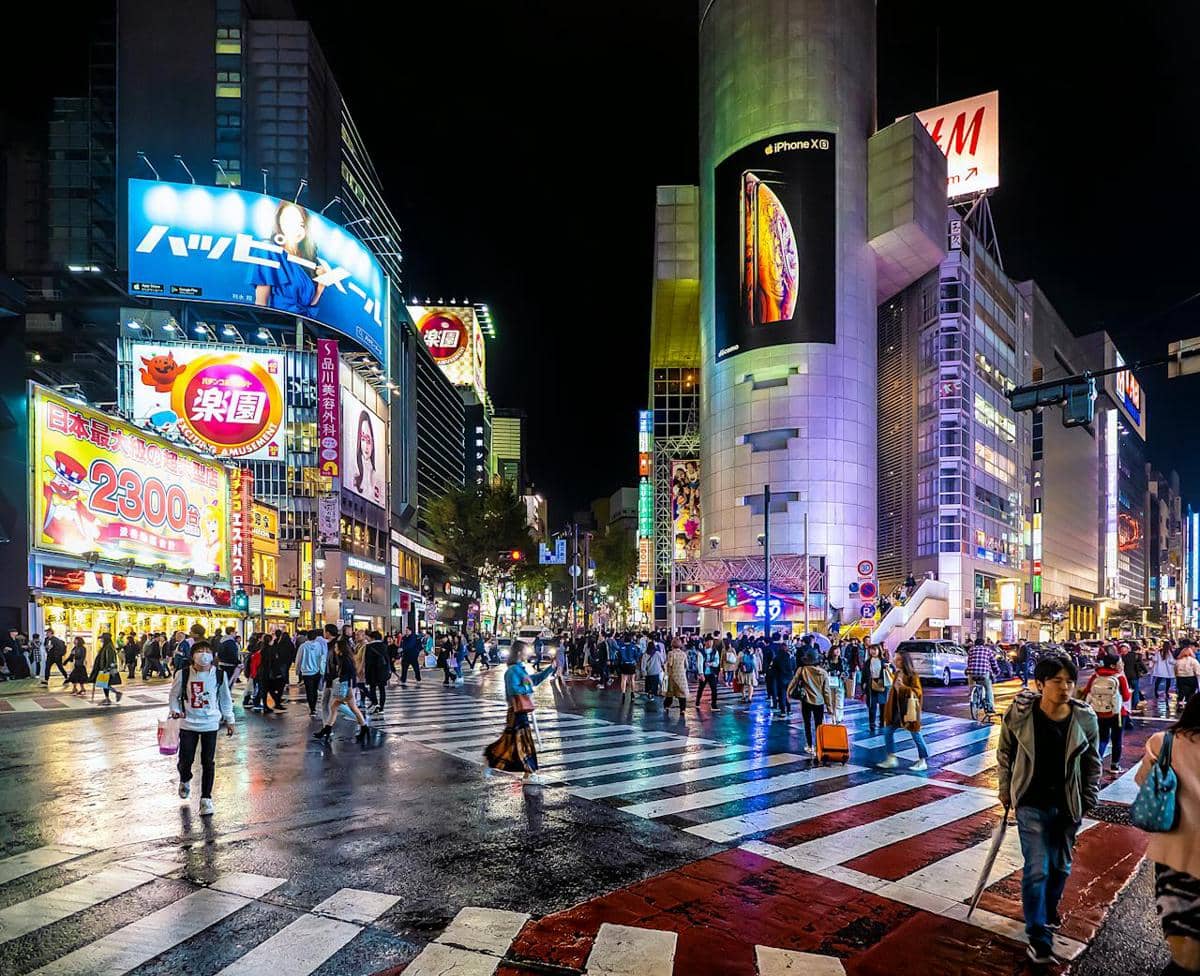 Keramaian malam di persimpangan Shibuya Tokyo dengan pejalan kaki menyeberang dan papan reklame warna-warni yang menyala terang.
