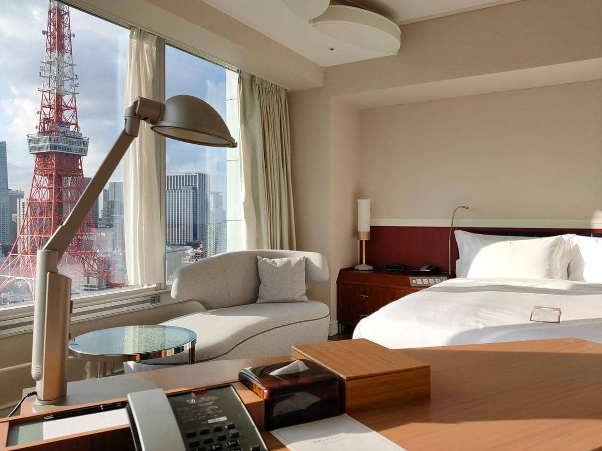 Kamar hotel modern dengan tempat tidur putih, meja kerja, dan pemandangan menara merah serta gedung-gedung kota dari jendela besar.