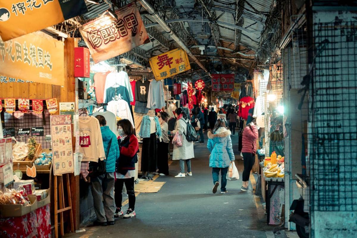 Suasana pertokoan di pasar malam dengan deretan kios menjual pakaian, makanan, dan berbagai barang, serta pengunjung yang berjalan di lorong sempit.