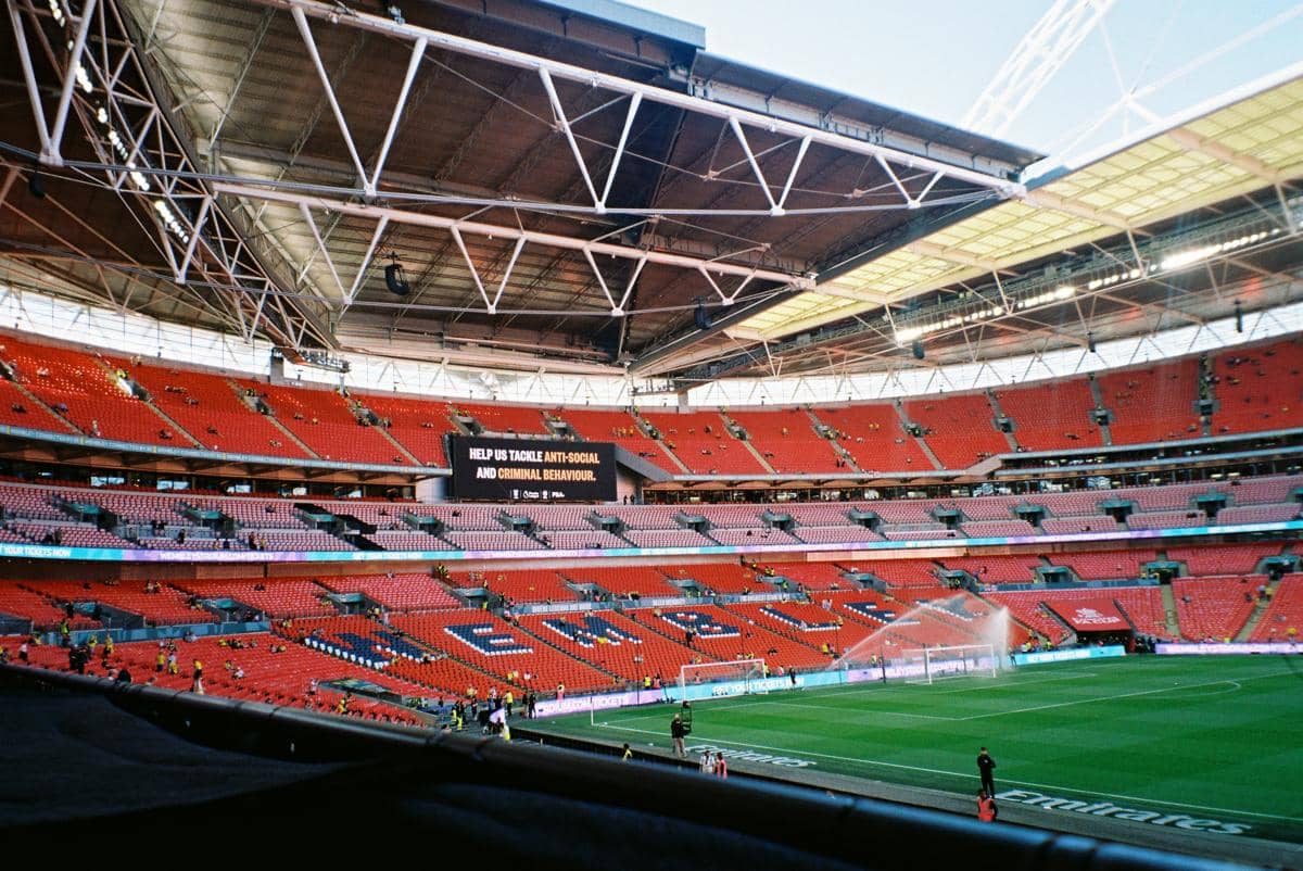 Pemandangan dalam stadion sepak bola besar dengan kursi tribun merah kosong, struktur atap besi yang rumit, dan alat penyiram air yang sedang membasahi rumput lapangan hijau.