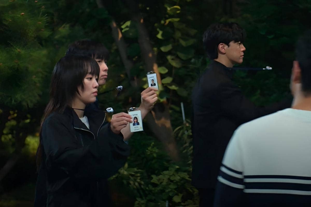 Beberapa karakter dalam drama Teach You a Lesson memegang kartu identitas di area luar ruangan yang dikelilingi pepohonan.